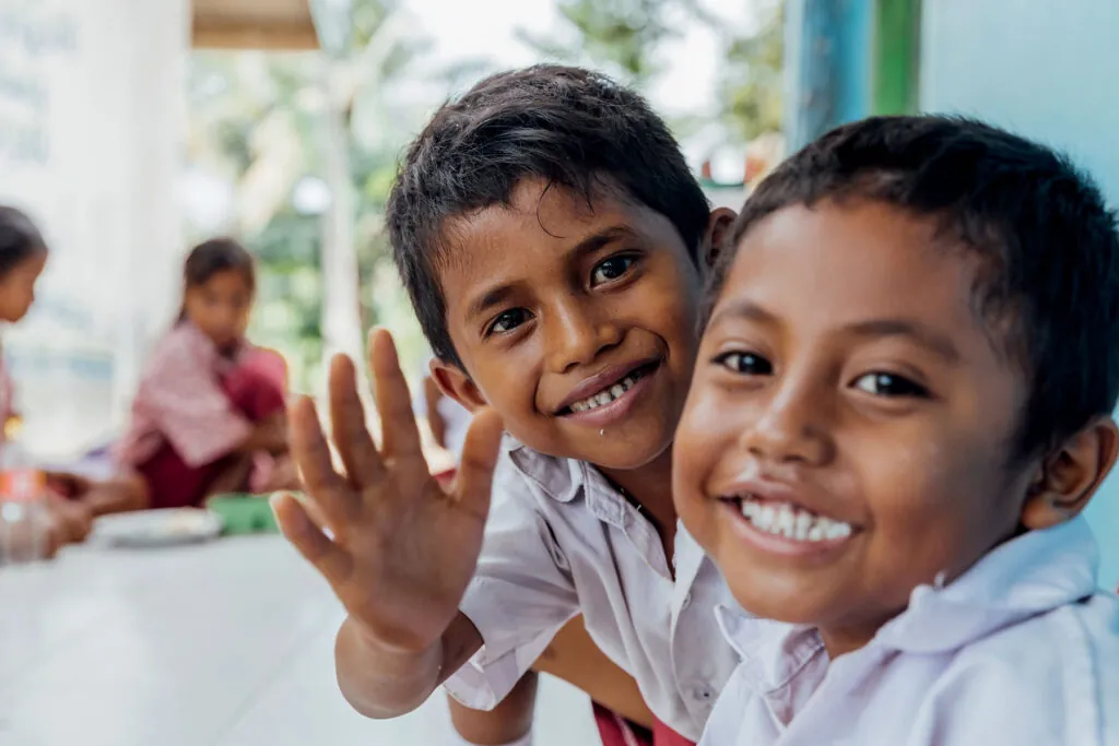 Two young Sumbanese boys in school uniforms smiling and waving during a classroom lunch session, reflecting the positive impact of educational programs supported by the Sumba Foundation and Burch Family Foundation for local communities.